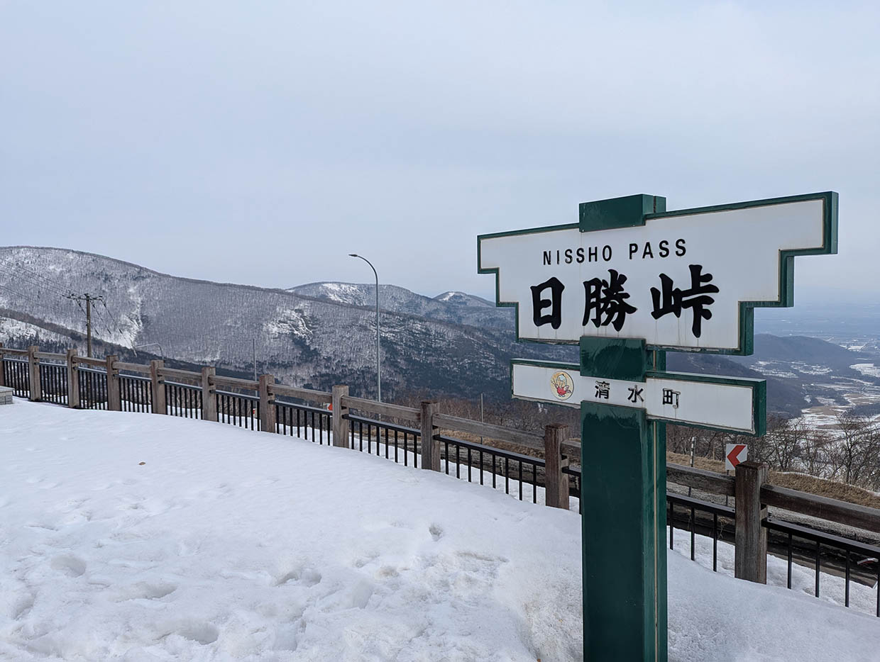 雪に覆われた日勝峠の展望台に立つ標識と、峠の向こうに広がる十勝平野の眺め。旅の終わりに立ち寄った峠で、道のりを振り返るひととき。