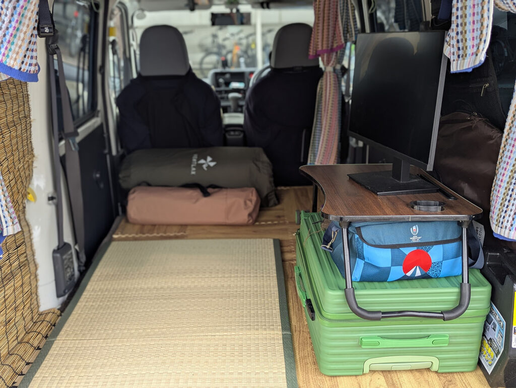 車中泊仕様の軽バンの車内に寝袋、畳、モニター、テーブルが整然と配置された様子。
