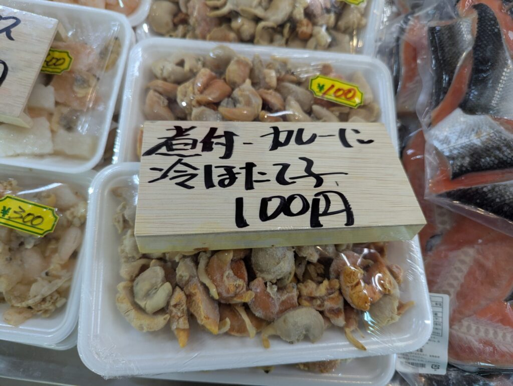 カットされたホタテがパックに詰められた冷凍食品。木札には『煮付・カレーに 冷ほたて子 100円』と手書きされている。周囲には他の海鮮パックも並び、市場の一角を感じさせる。