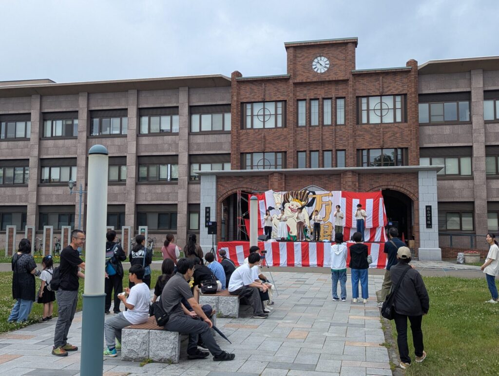 帯広畜産大学のレンガ造りの校舎前に設置された特設ステージ。赤白幕に囲まれた舞台では学生たちがパフォーマンスを行っており、観客がベンチや芝生に座って見守っている。