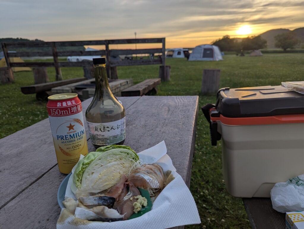達古武キャンプ場のテーブルで、ノンアルコールビールと焼酎の瓶、生キャベツや魚の刺身を並べて夕暮れの晩酌を楽しむ様子。