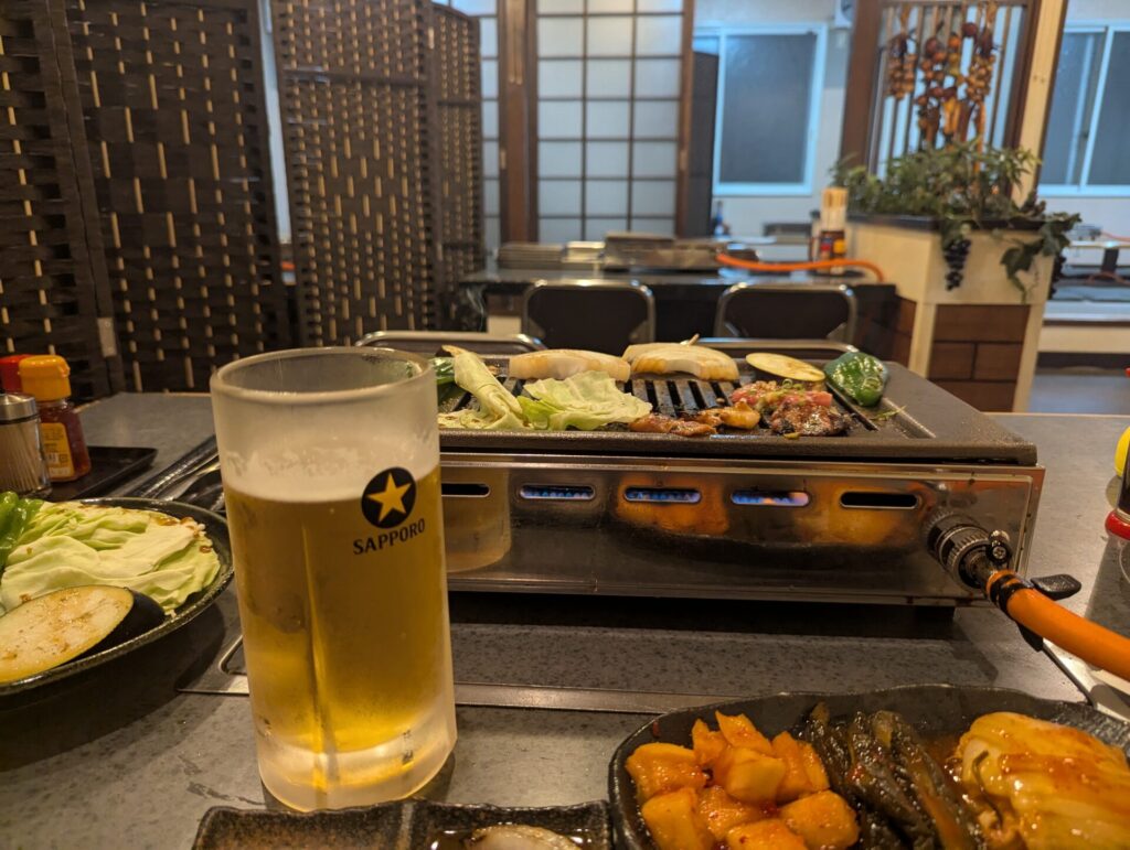 焼肉の煙が立ちのぼるテーブルに、冷えたサッポロ生ビールが置かれている。奥には網の上の肉と野菜が並び、居心地の良い店内の様子も映る。