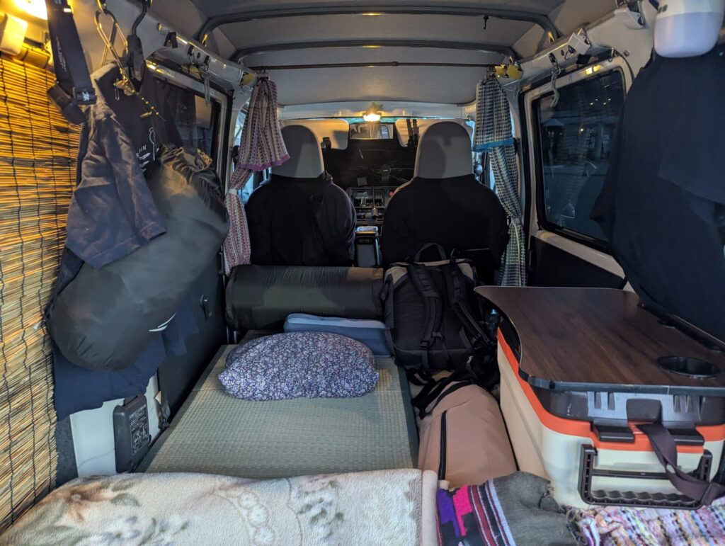 車内に敷いた寝具と枕、簡易テーブルや荷物が整然と並び、カーテンで仕切られた空間が旅人の寝床になった車中泊の様子。小さな空間に工夫が詰まった快適な寝床が広がる。