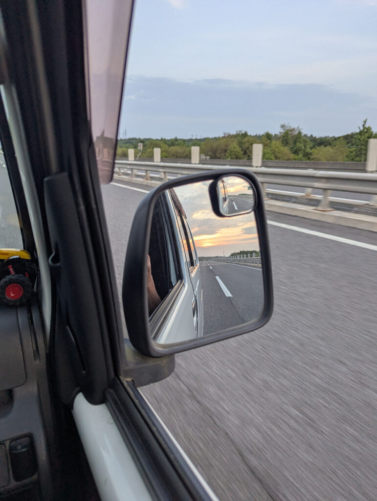 車のサイドミラーに映る夕焼けと、高速道路を走る車の車窓