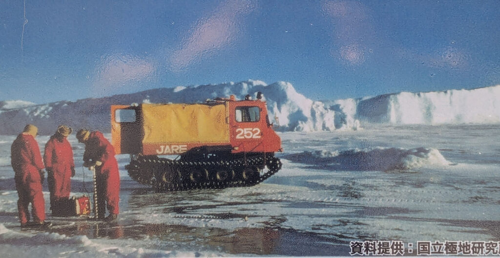 南極の氷上で作業する赤い防寒服の隊員たちと、雪上車SM252。背景には氷壁が広がる。