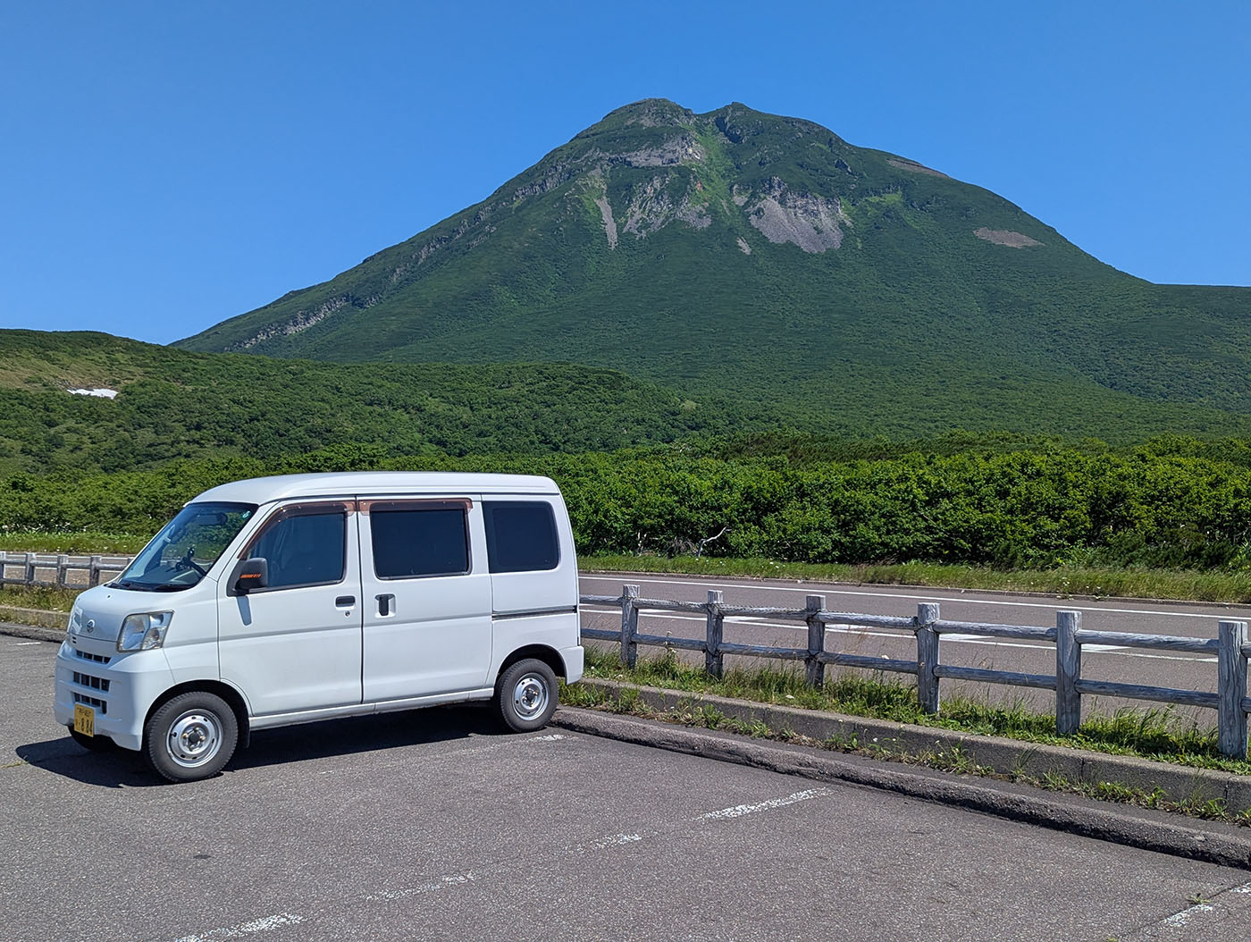 広大な緑の山を背景に、駐車場に停まる白い軽バン。快晴の青空が広がる北海道の風景。