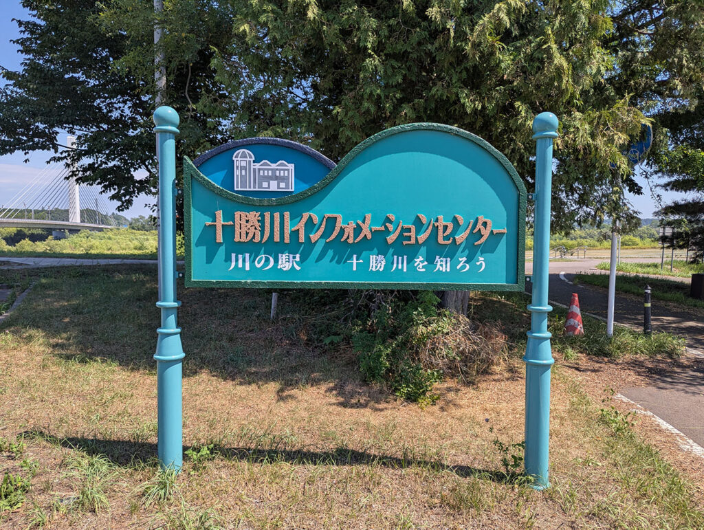 十勝川インフォメーションセンターの緑色の看板と背景に見える吊り橋と緑の木々。