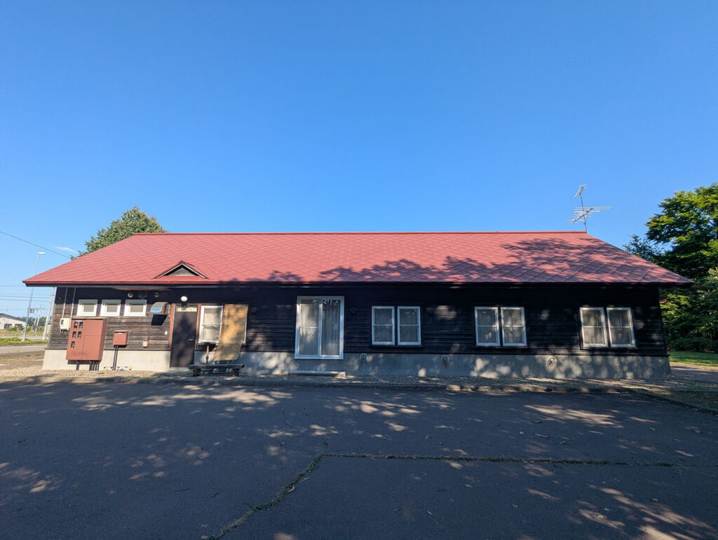 赤い屋根と黒い外壁の旧駅逓再現建物を裏側から撮影。青空に建物のシルエットがくっきり浮かぶ。