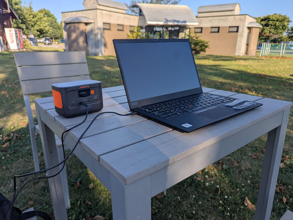 芝生に置かれた白いテーブルで、ポータブル電源とノートパソコンを使って作業する様子。道の駅しかおいの広々とした空間が背景に映る。