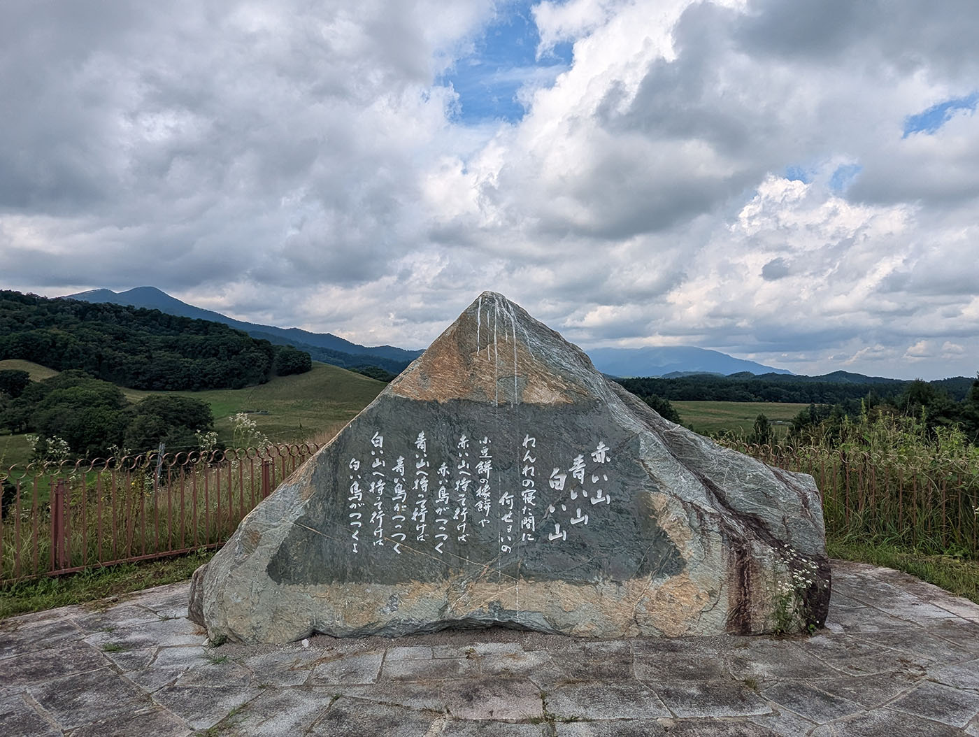 八千代牧場に建つ石碑。山並みと広大な牧草地を背景に、「赤い山 青い山 白い山」と刻まれた詩が大きな岩に刻まれている。