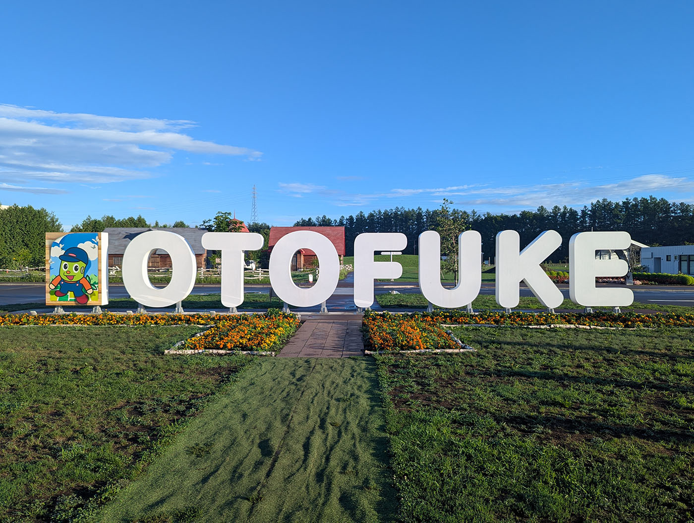 道の駅おとふけの入口にある「OTOFUKE」の大型モニュメントとマスコットキャラクターのパネル。晴天の下、花壇の花が彩りを添えている。