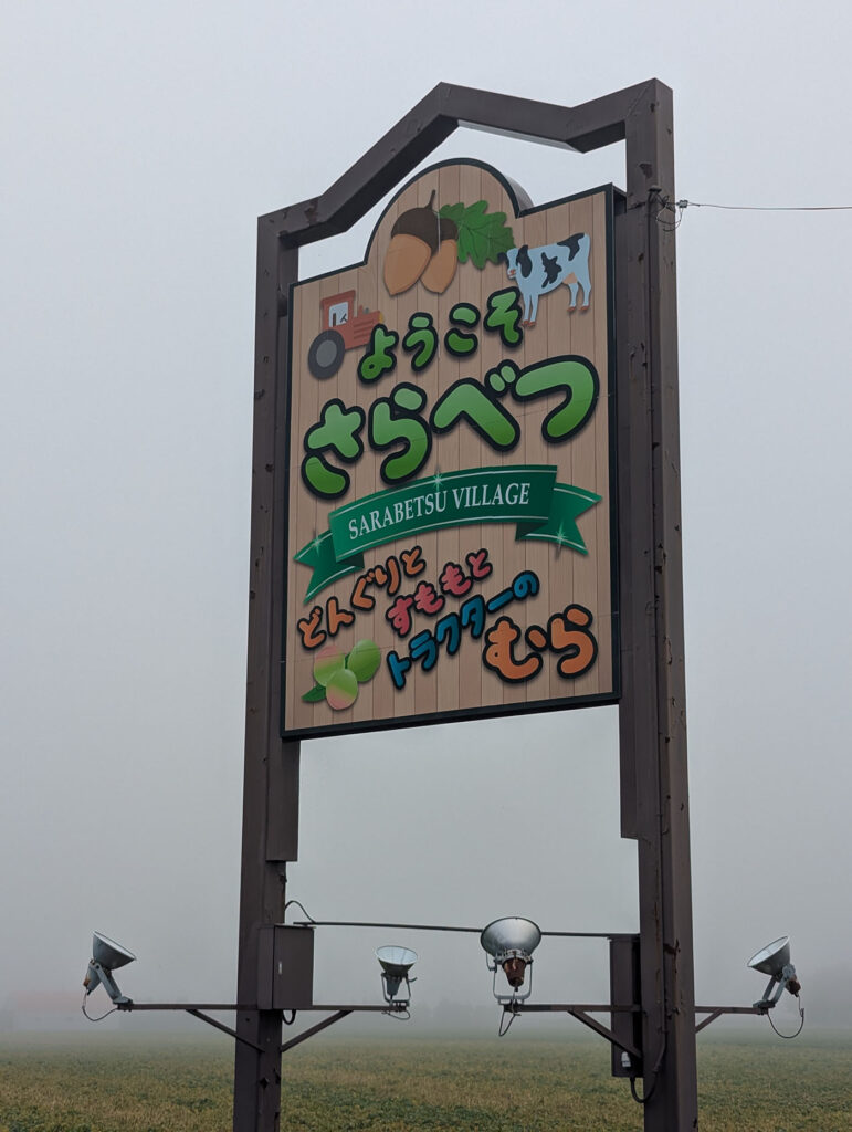 霧の中に立つ「ようこそ さらべつ」の看板。どんぐりや牛、すもも、トラクターのイラストが描かれている。