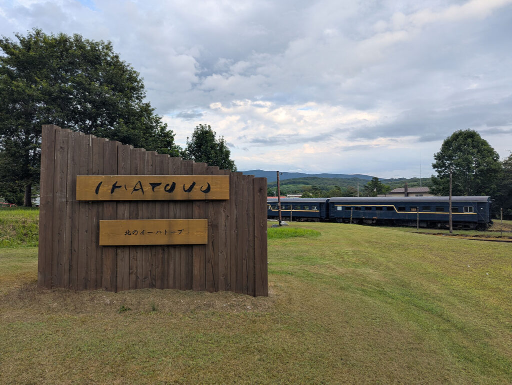 北海道むかわ町の富内銀河ステーション入口に立つ木製の看板「IHATOV 北のイーハトーブ」と、背景に並ぶ保存客車。廃線跡の広場に青い車両が静かに佇む。
