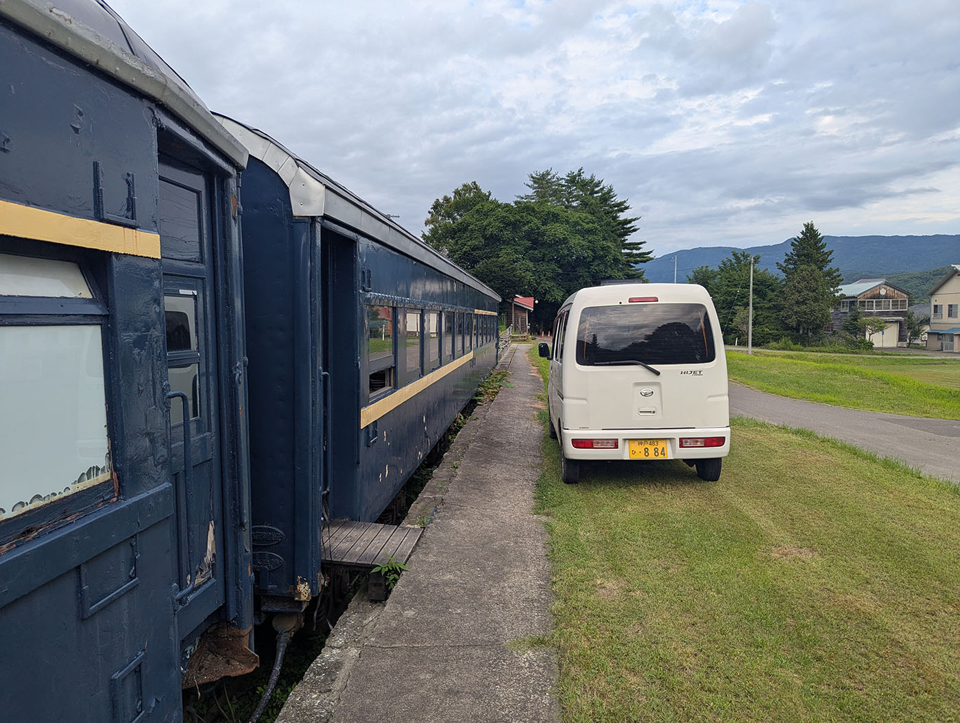 北海道むかわ町の富内銀河ステーション。保存された青い客車と、隣に駐車する白い軽バン。背景には緑の芝生と山並みが広がる。