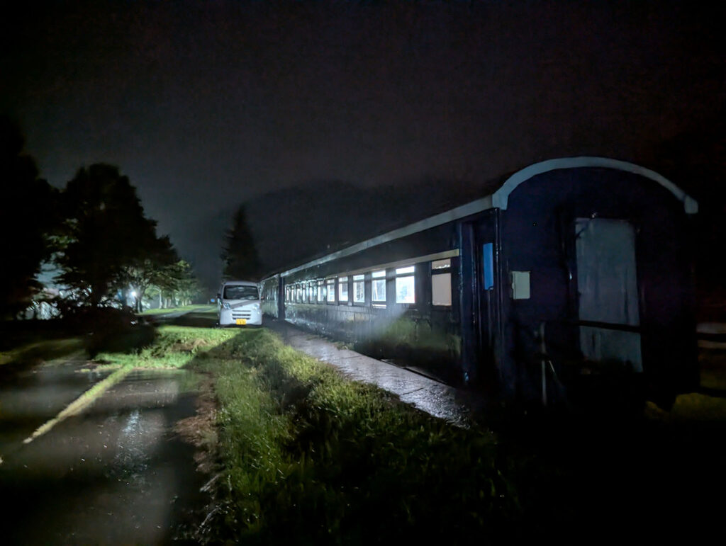 夜の闇の中、車内に灯りがともった古い客車が静かに停まっている。外は雨で、列車の窓明かりだけが周囲を柔らかく照らしている。