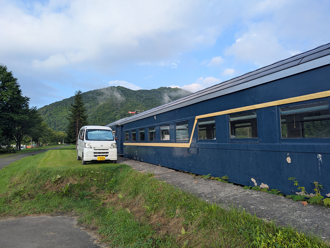 富内銀河ステーションのホームに停められた白い軽バンと、青色の旧客車。背後には夏の緑に覆われた山並みが広がり、快晴の朝の静けさを感じさせる。