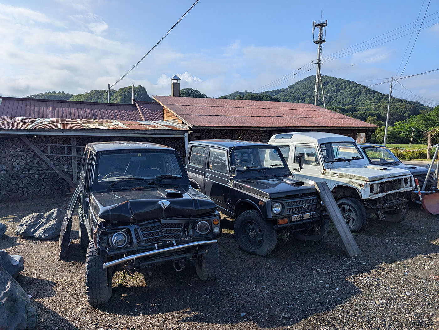 錆びたトタン屋根の前に並ぶ3台のスズキ・ジムニー。黒、黒、白の順で、いずれも長年の風雪に耐えた風