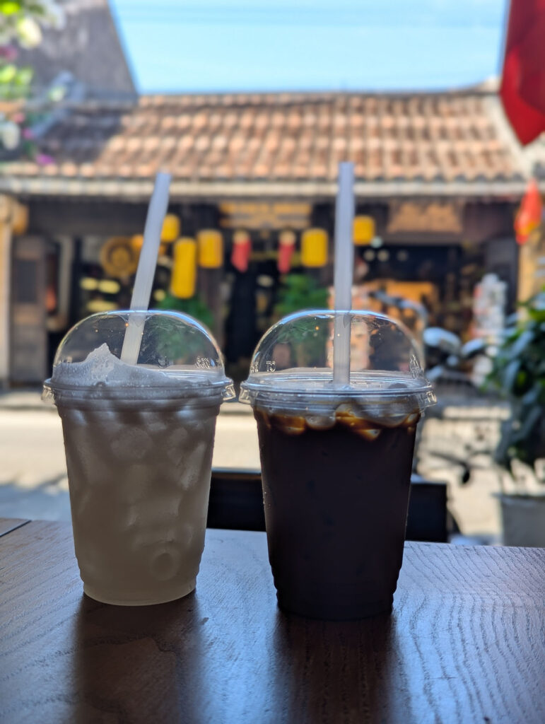 ホイアン旧市街のカフェ「92ステーション」で頼んだアイスコーヒーとライムジュース。木のテーブルに並び、背景にはランタンが吊るされた街並みがぼんやり映っている。