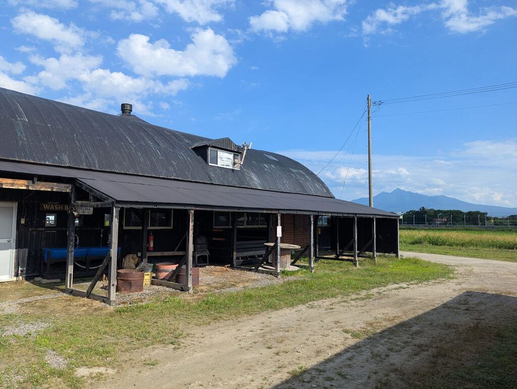 黒い屋根の大きな建物と、その奥に広がる青空と知床連山。かつての牛舎を活用したクリオネキャンプ場の一部。