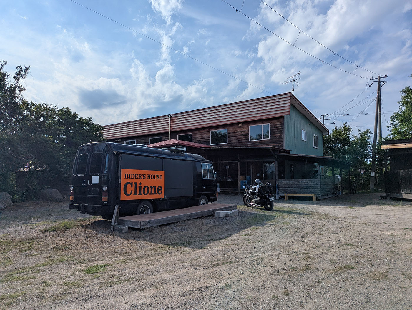 「RIDER'S HOUSE Clione」と書かれた黒いバスと、奥にある木造の管理棟。ライダーの旅拠点として人気のキャンプ場。