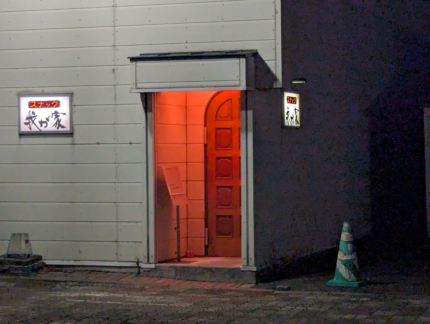 夜の街角に佇む「スナック我が家」の入り口。赤い照明が漏れ、静かな通りに看板が光っている。