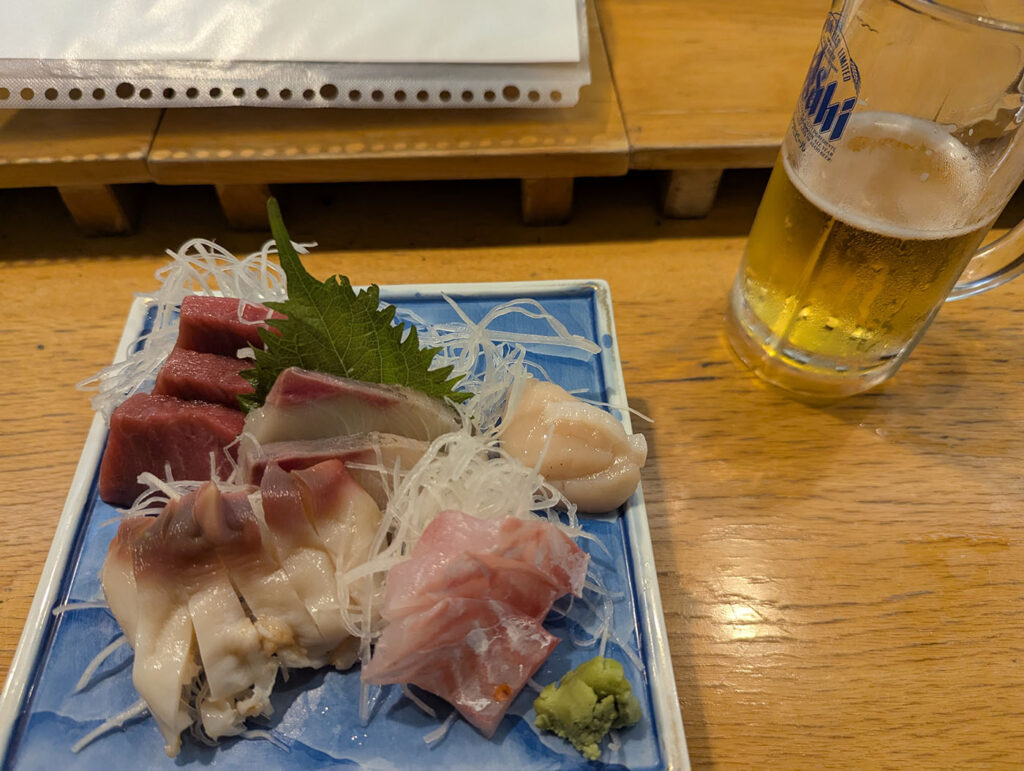 びっくり寿司で提供された新鮮な刺身盛り合わせとグラスビール。木のカウンター席で撮影された一枚。