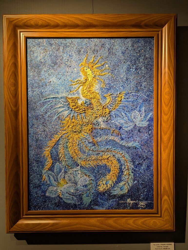 青を基調に、金色の鳳凰が花とともに舞う姿を描いた絵画。厚みのある質感と鮮やかなコントラストが特徴的。