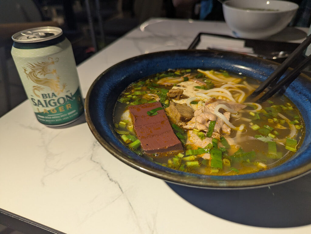 フエ地方のフォー（ブンボーフエ）の一杯と、サイゴンビールの缶。濃いスープと具材が特徴的な麺料理。