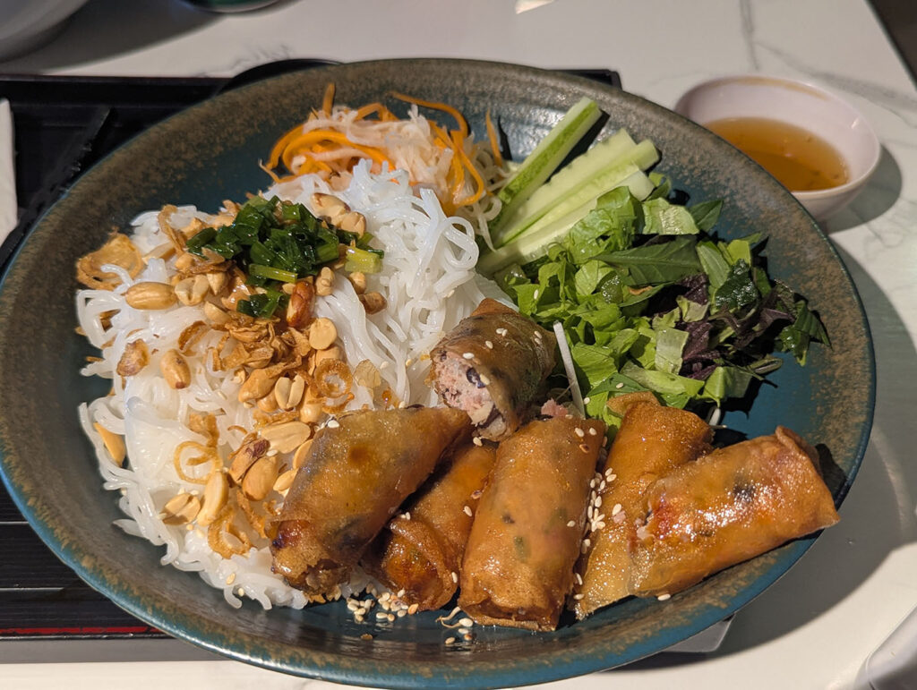揚げ春巻きと香草、ブン（米麺）が盛り付けられたベトナム料理の一皿。ナッツやネギ油がトッピングされ、ヌクチャムの小皿も添られている。