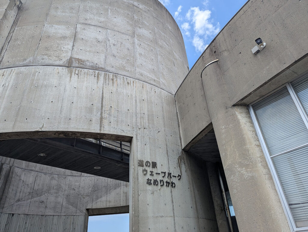 道の駅ウェーブパークなめりかわのコンクリート建築のディテール。円筒形の塔と曲線を描く壁面に、「道の駅 ウェーブパーク なめりかわ」の文字が刻まれている。
