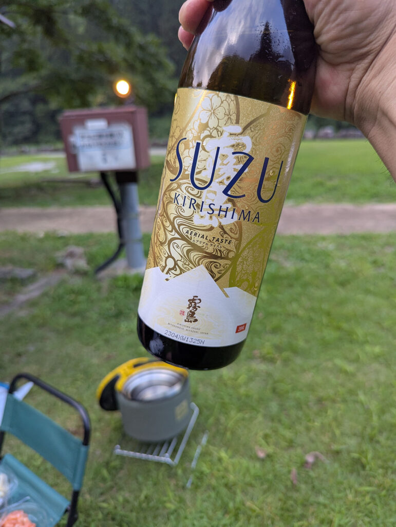 片貝山ノ守キャンプ場で「SUZU KIRISHIMA」のボトルを掲げる手元。緑の芝生と夕暮れのランタンが背景に映る。