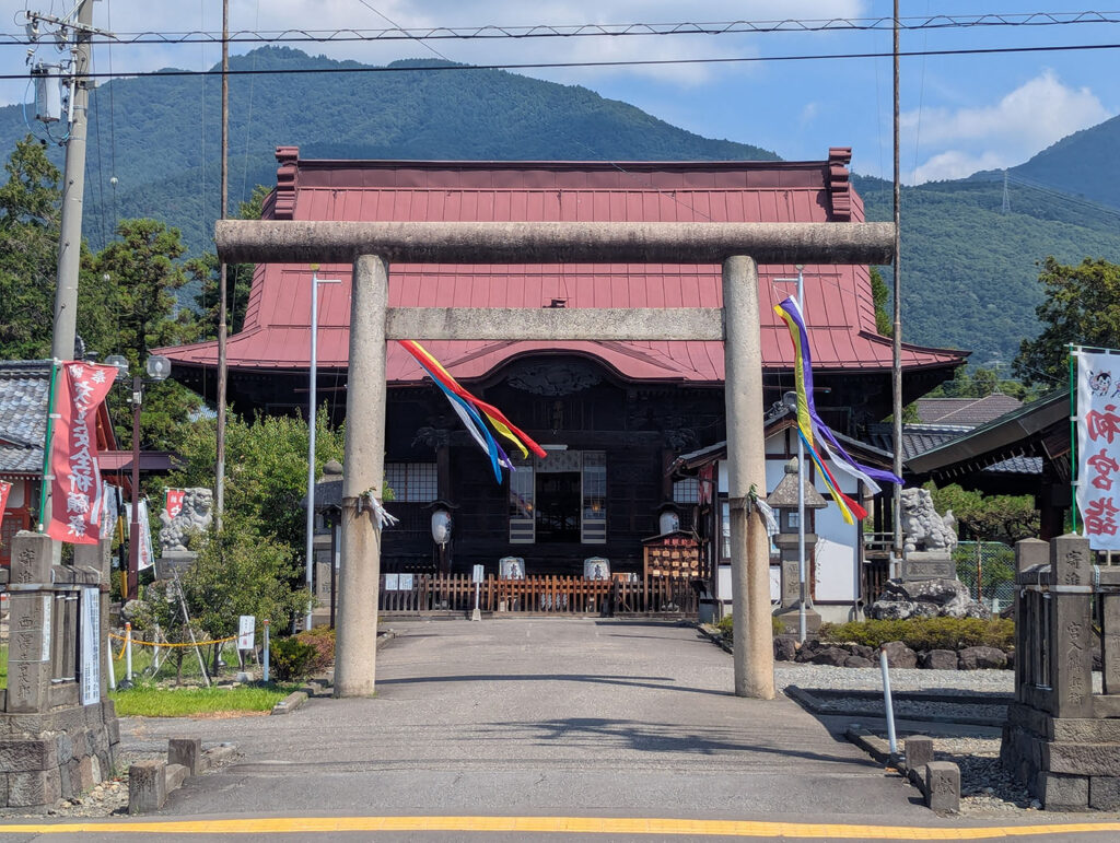 長野県上田市の上田大神宮の正面。石の鳥居の向こうに赤い屋根の本殿があり、五色の吹き流しが風になびいている。背後には緑濃い山々がそびえる。