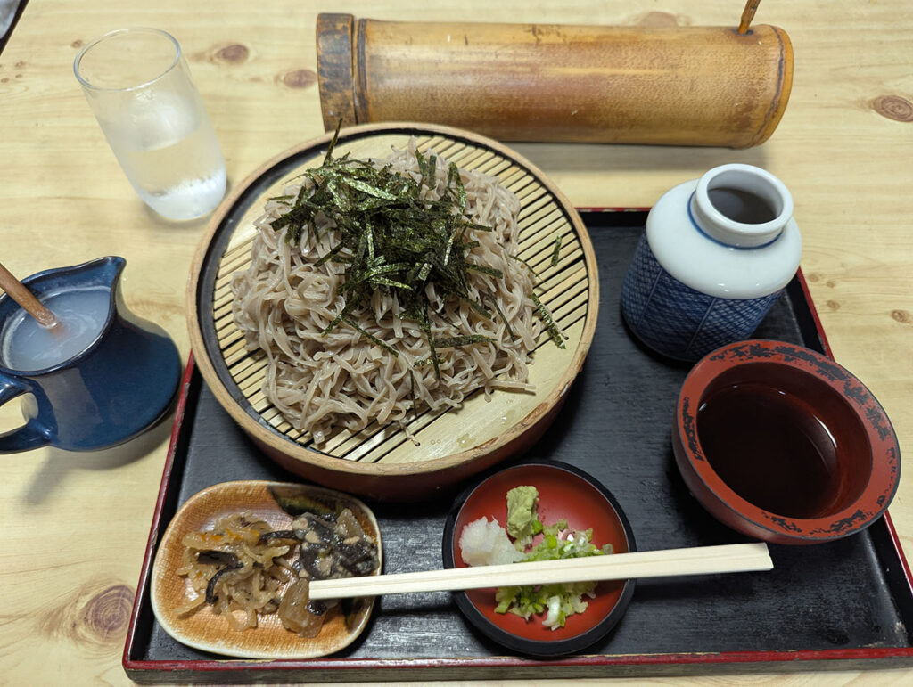 長野県上田市・塩田のそば屋で出されたざるそばの膳。細く短いそばに刻み海苔がのり、つけ汁・薬味・そば湯・小鉢が添えられている。竹筒の箸入れが風情を添える。