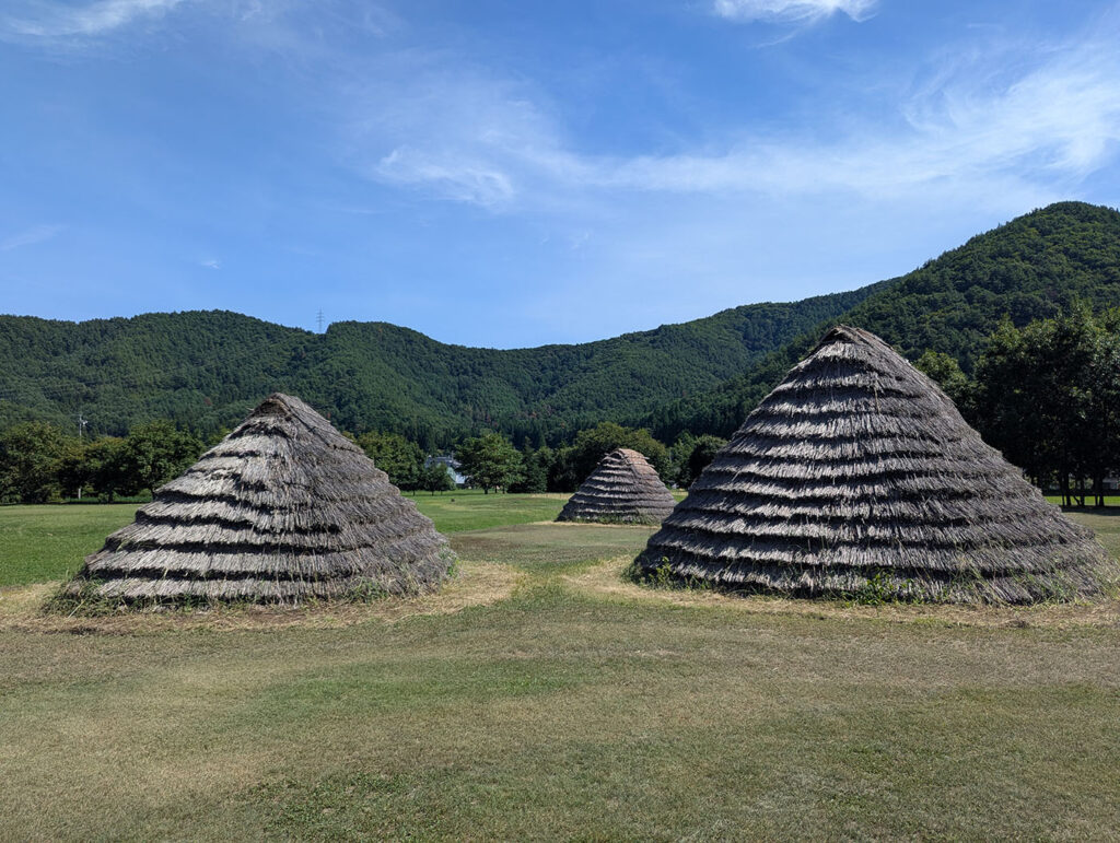 長野県塩尻市の平出遺跡に復元された竪穴住居群。円錐形の茅葺き屋根が広い草地に点在し、背後には緑濃い山並みが広がる。