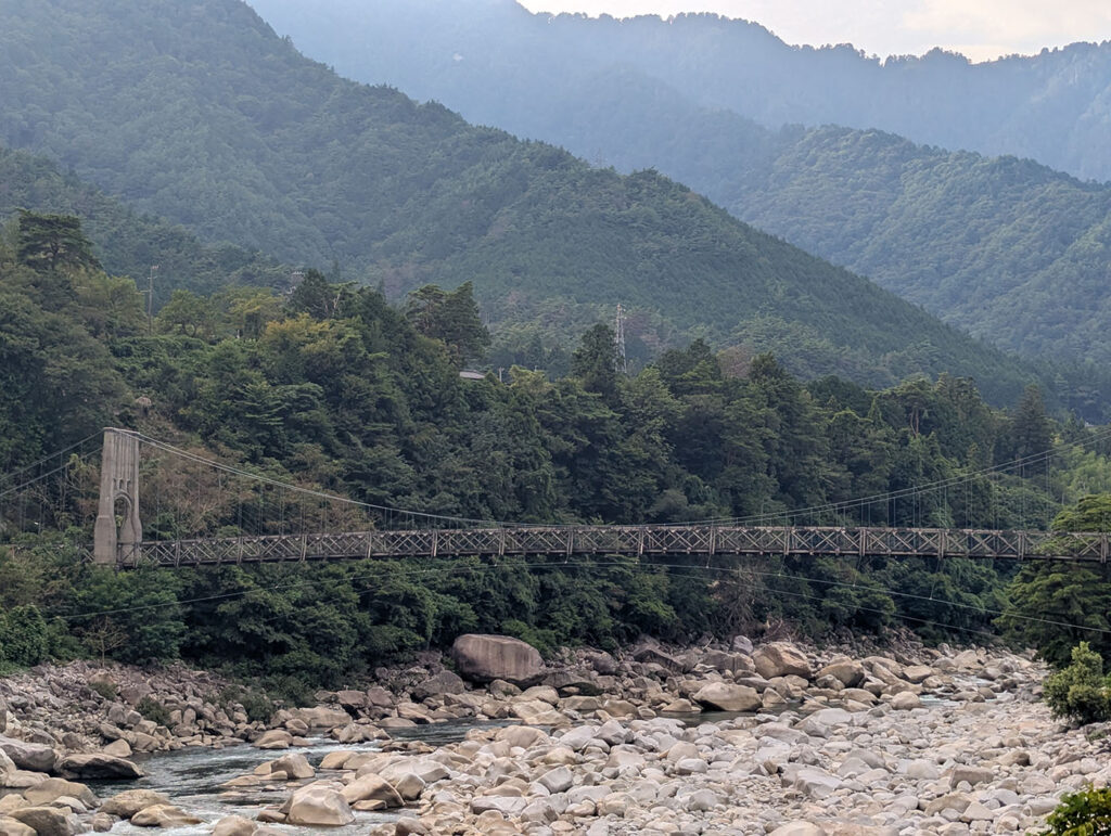 南木曽町の山あいにかかる桃介橋。木製トラスの吊り橋が、岩の多い木曽川をまたぎ、背後には幾重にも重なる山々が広がっている。